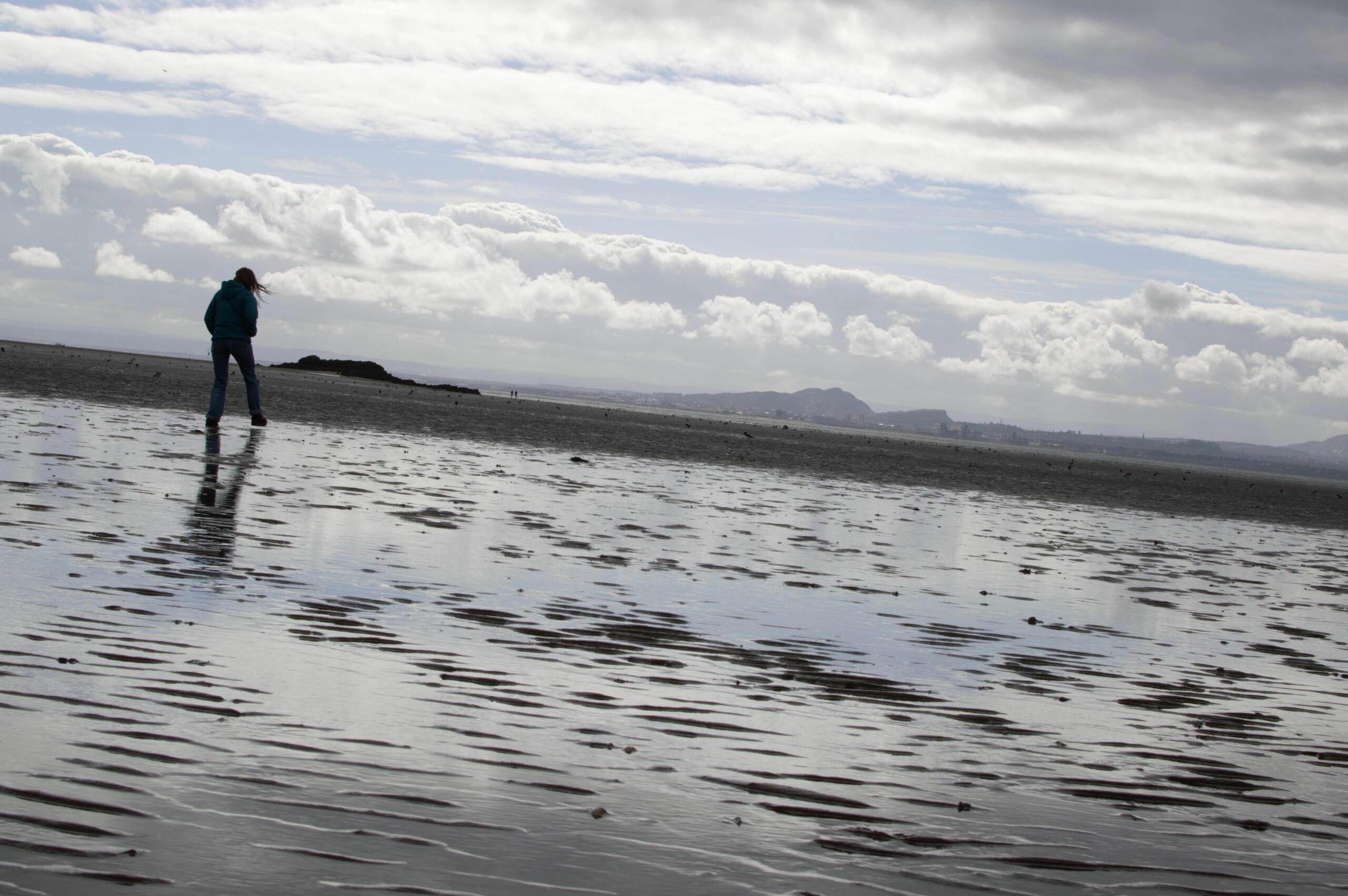 Photograph of a beach and a grey sky with a figure taking a step in the foreground and the skyline of Edinburgh in the background, with the horizon at a slight tilt